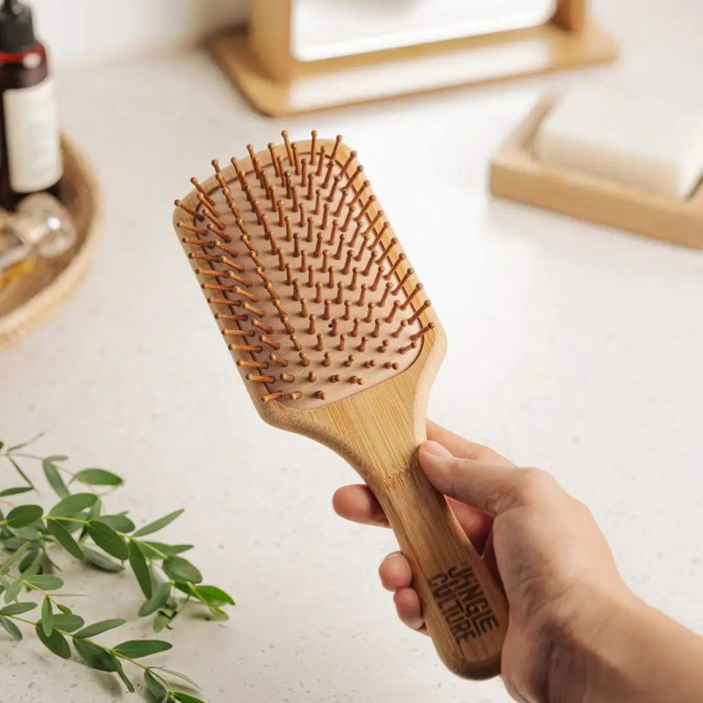 A person's hand is holding a bamboo hairbrush with wooden bristles, indicating the product's ergonomic design for comfortable use in daily hair care. The background includes a neutral-toned bathroom setting, suggesting the brush's place in an eco-friendly lifestyle.
