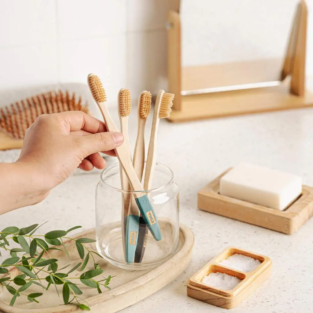 A hand selects a bamboo toothbrush from a glass jar on a bathroom countertop, showcasing sustainable dental hygiene products.