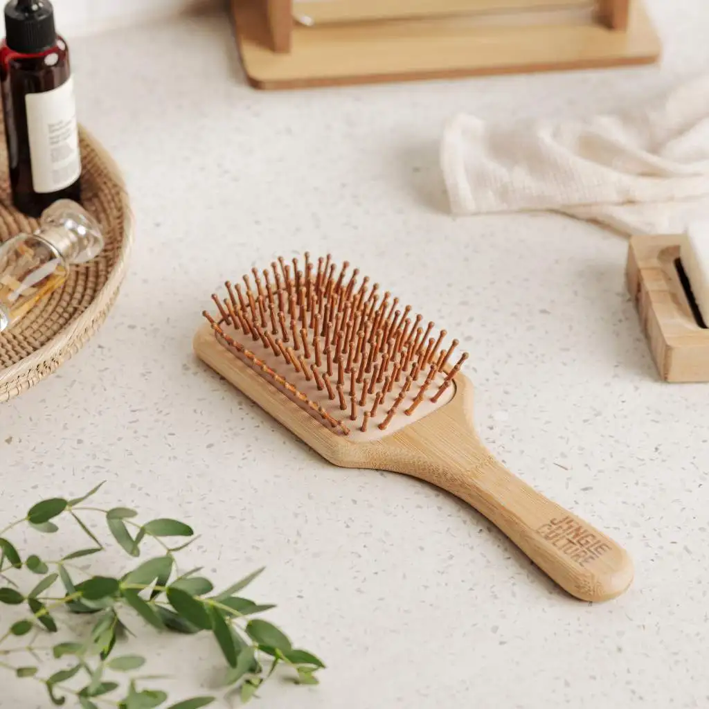 A single bamboo hairbrush with natural wooden bristles lies on a minimalist, lightly speckled surface, next to a glass bottle and some green foliage, highlighting a sustainable beauty routine.
