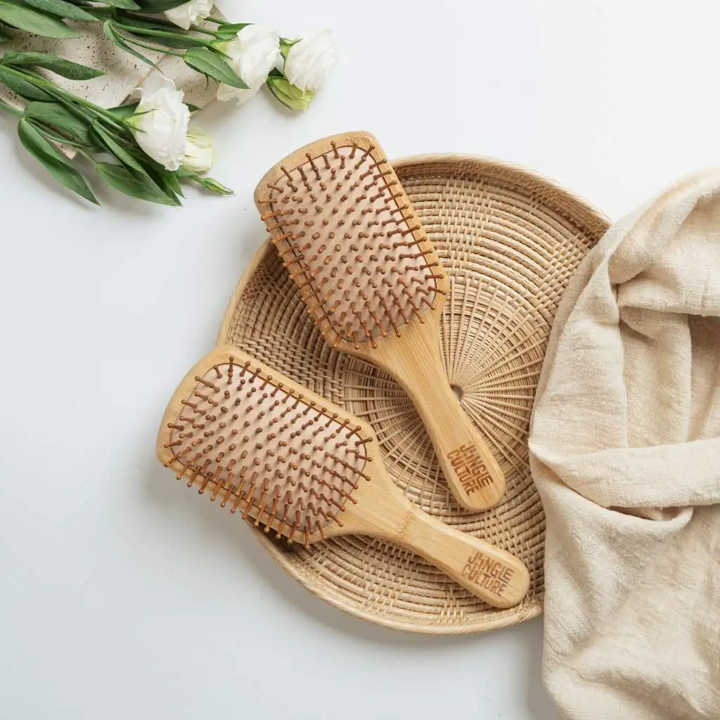 Two wooden hairbrushes with bamboo bristles are displayed on a woven rattan tray, accompanied by white flowers and a beige cloth, suggesting an eco-conscious grooming experience.